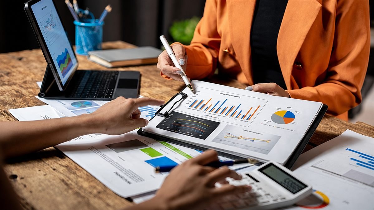 Dos personas revisando gráficos comparativos y tableros de indicadores impresos sobre una mesa de madera, con una tableta y una calculadora al alcance